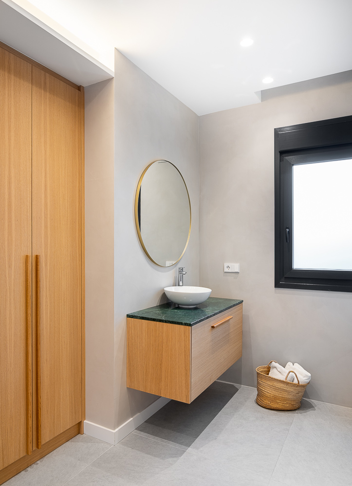 Aseo con espejo redondo dorado y mueble de madera suspendido con lavabo sobre encimera, interiorismo de Arquitectura de la Ribera