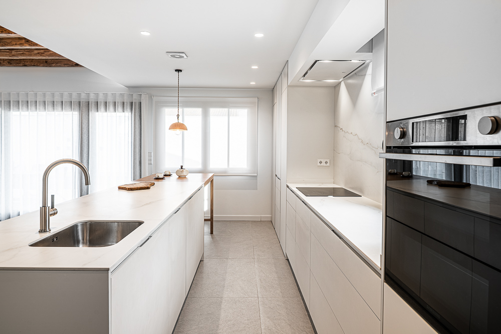 Cocina en blanco con fregadero bajo ventana y columna de hornos negros, proyecto de interiorismo en Corella