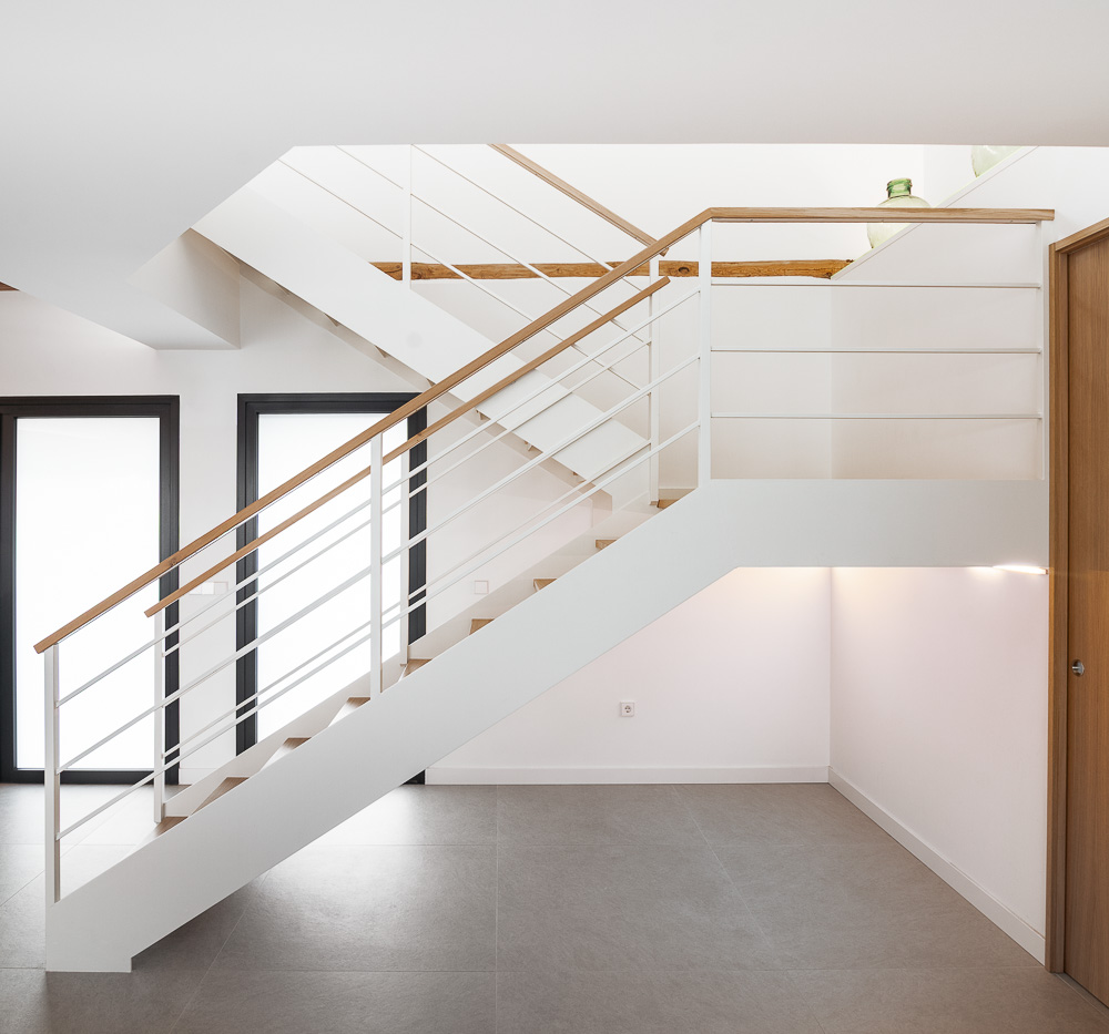 Escalera blanca con peldaños de madera en doble altura bajo vigas vistas, arquitectura de vivienda unifamiliar en Navarra