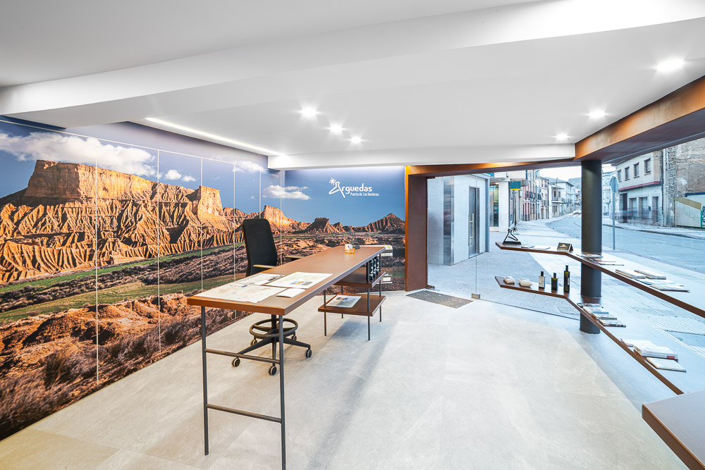 Mostrador de madera junto a fachada de vidrio y mural de las Bardenas, interiorismo de oficina de turismo en Arguedas